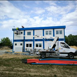 PODNOŚNIKI CEZAR CEZARY BIEDRZYCKI - Montaż modułowego budynku biurowego w Jadwisinie z użyciem podnośnika koszowego; widoczni dwaj pracownicy na dachu i rury instalacyjne na pierwszym planie.