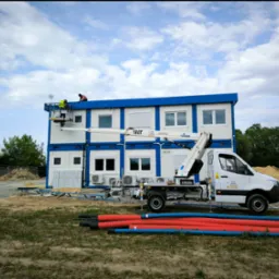 Montaż modułowego budynku biurowego w Jadwisinie z użyciem podnośnika koszowego; widoczni dwaj pracownicy na dachu i rury instalacyjne na pierwszym planie.