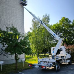 PODNOŚNIKI CEZAR CEZARY BIEDRZYCKI - Dwóch pracowników na podnośniku koszowym, w żółtych kamizelkach, przy ścianie budynku z cegły, prawdopodobnie podczas prac konserwacyjnych lub naprawczych elewacji.