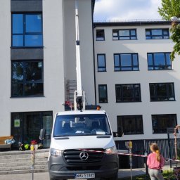 PODNOŚNIKI CEZAR CEZARY BIEDRZYCKI - Pracownik na podnośniku koszowym przy białej elewacji budynku z dużymi oknami, biały samochód Mercedes z przodu, zabezpieczony teren, osoba obserwująca z boku.