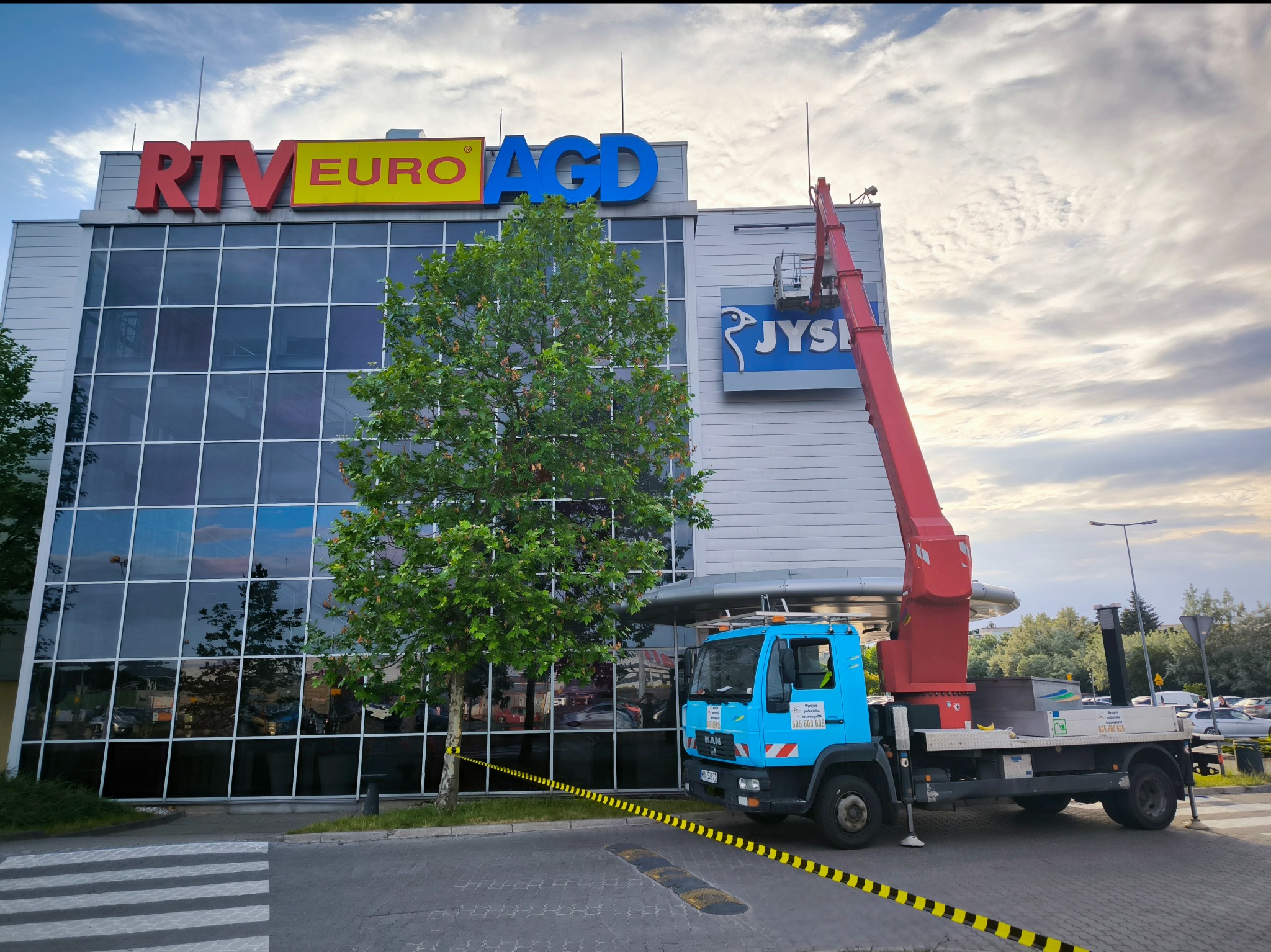 Ciężarówka z podnośnikiem koszowym na tle budynku RTV Euro AGD, osoba w koszu przy znaku JYSK, taśma ostrzegawcza wokół drzewa.