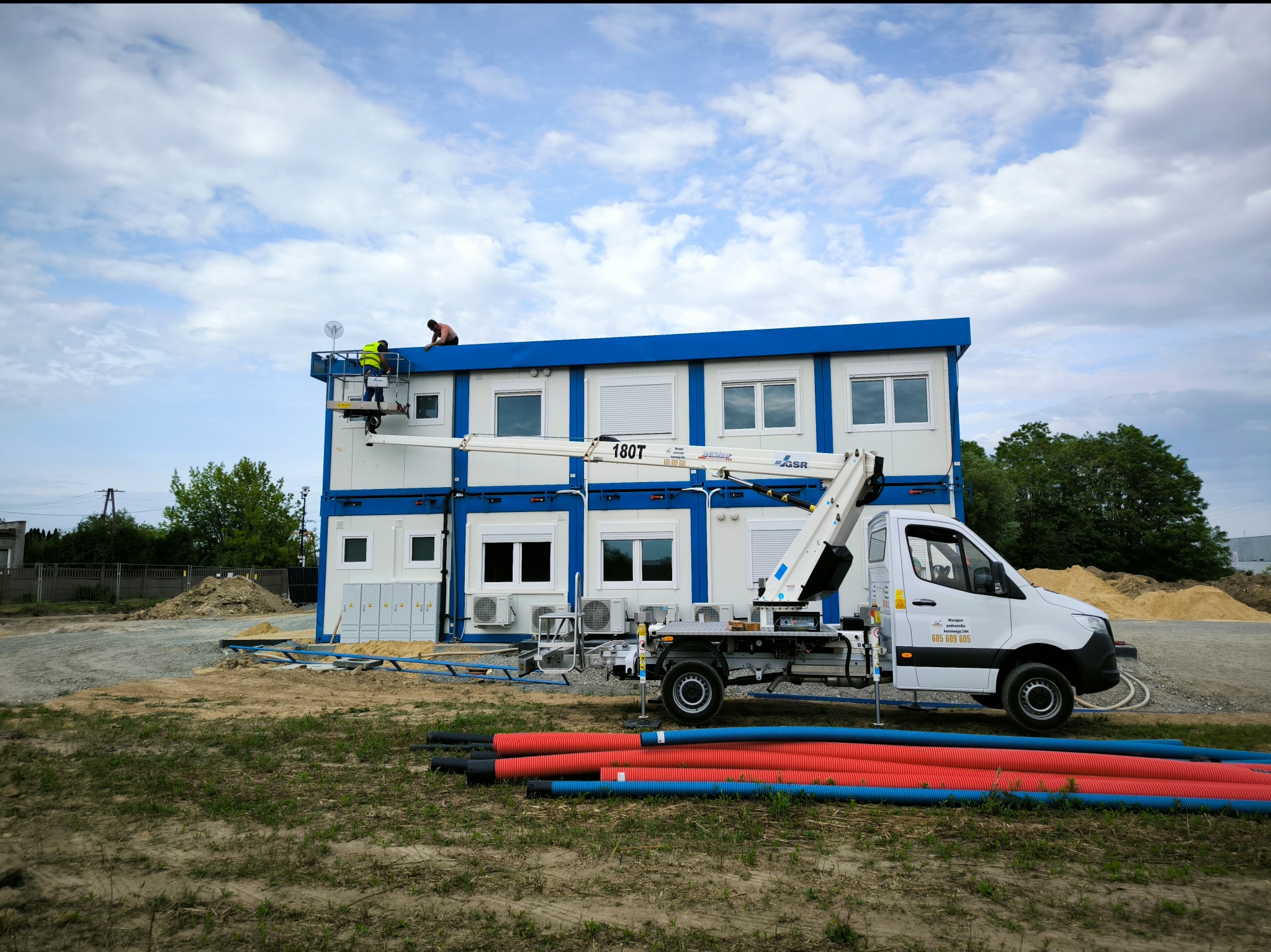 Montaż modułowego budynku biurowego w Jadwisinie z użyciem podnośnika koszowego; widoczni dwaj pracownicy na dachu i rury instalacyjne na pierwszym planie.