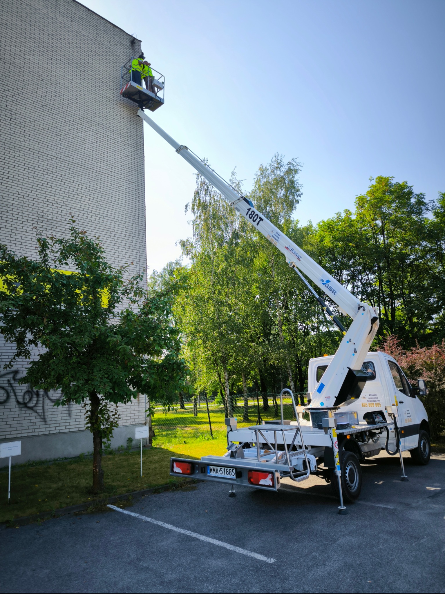 Dwóch pracowników na podnośniku koszowym, w żółtych kamizelkach, przy ścianie budynku z cegły, prawdopodobnie podczas prac konserwacyjnych lub naprawczych elewacji.