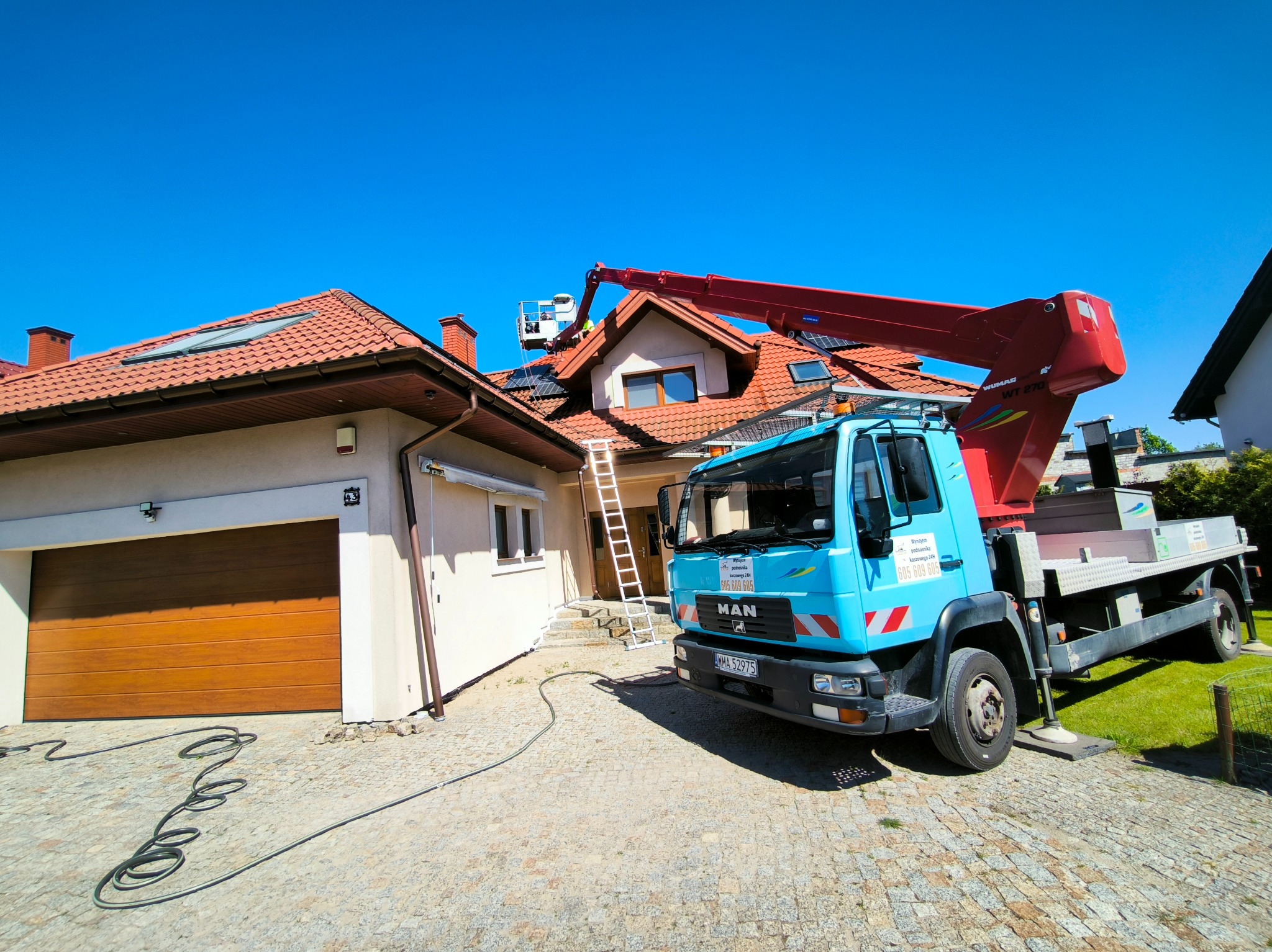 Praca na wysokościach: platforma koszowa na podwoziu samochodu MAN z wysięgnikiem ustawionym przy dachu domu jednorodzinnego z czerwoną dachówką, widoczna drabina oparta o dach i pracownik w koszu...