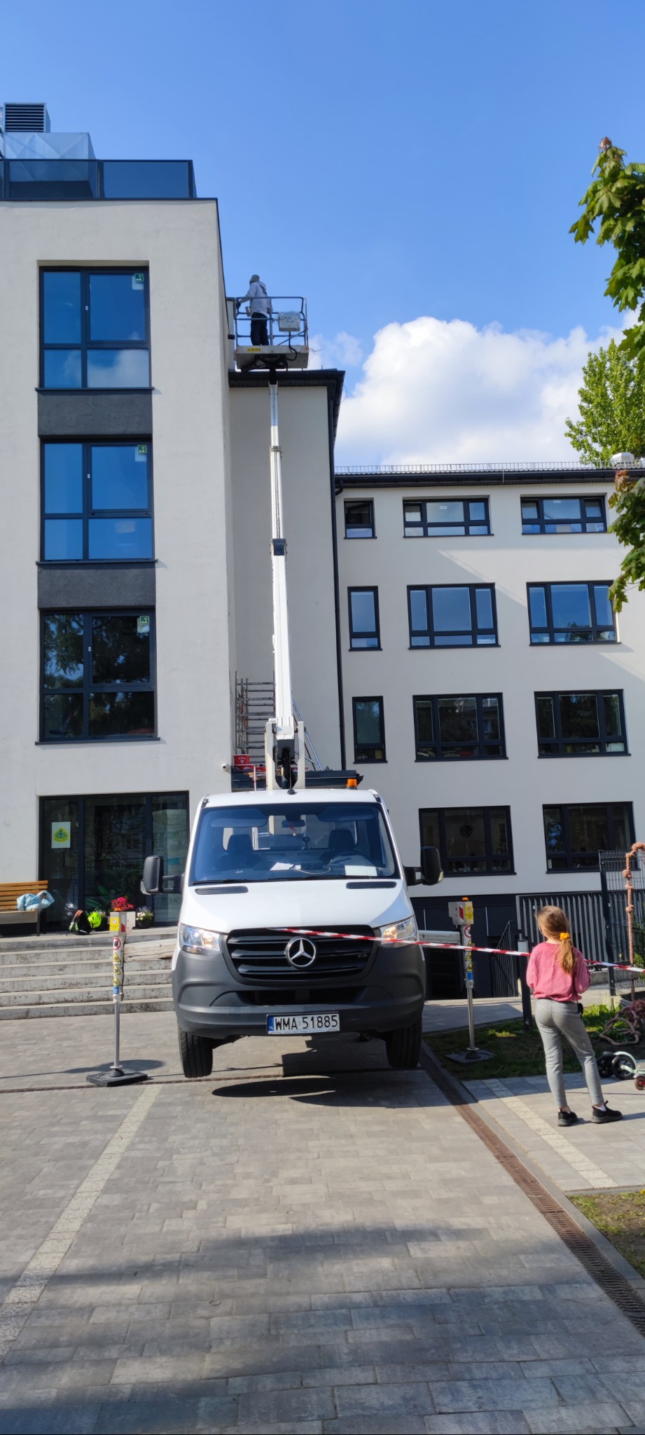 Pracownik na podnośniku koszowym przy białej elewacji budynku z dużymi oknami, biały samochód Mercedes z przodu, zabezpieczony teren, osoba obserwująca z boku.
