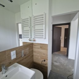 F.H.U.ZABRZESKI Rafał Zabrzeski - Wnętrze łazienki w trakcie wykańczania, widoczna umywalka, toaleta, grzejnik drabinkowy na ścianie z imitacją drewna, surowa podłoga i otwarte drzwi do kolejnego pomieszczenia.