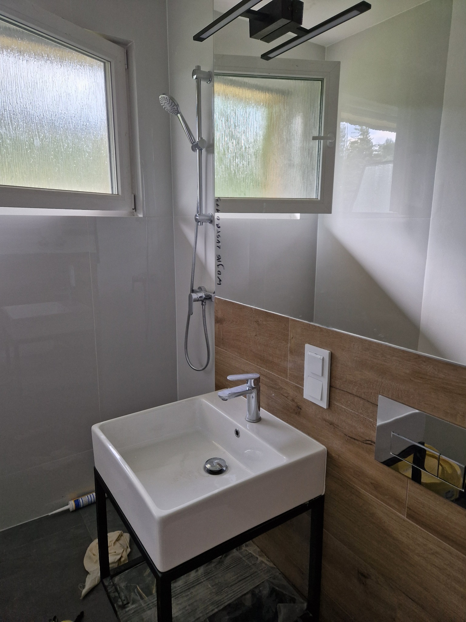 Nowoczesna łazienka z białą umywalką na czarnym stelażu, baterią, prysznicem i oknem z matową szybą, wykończona białymi i drewnopodobnymi płytkami, widoczna tuba silikonu na podłodze.