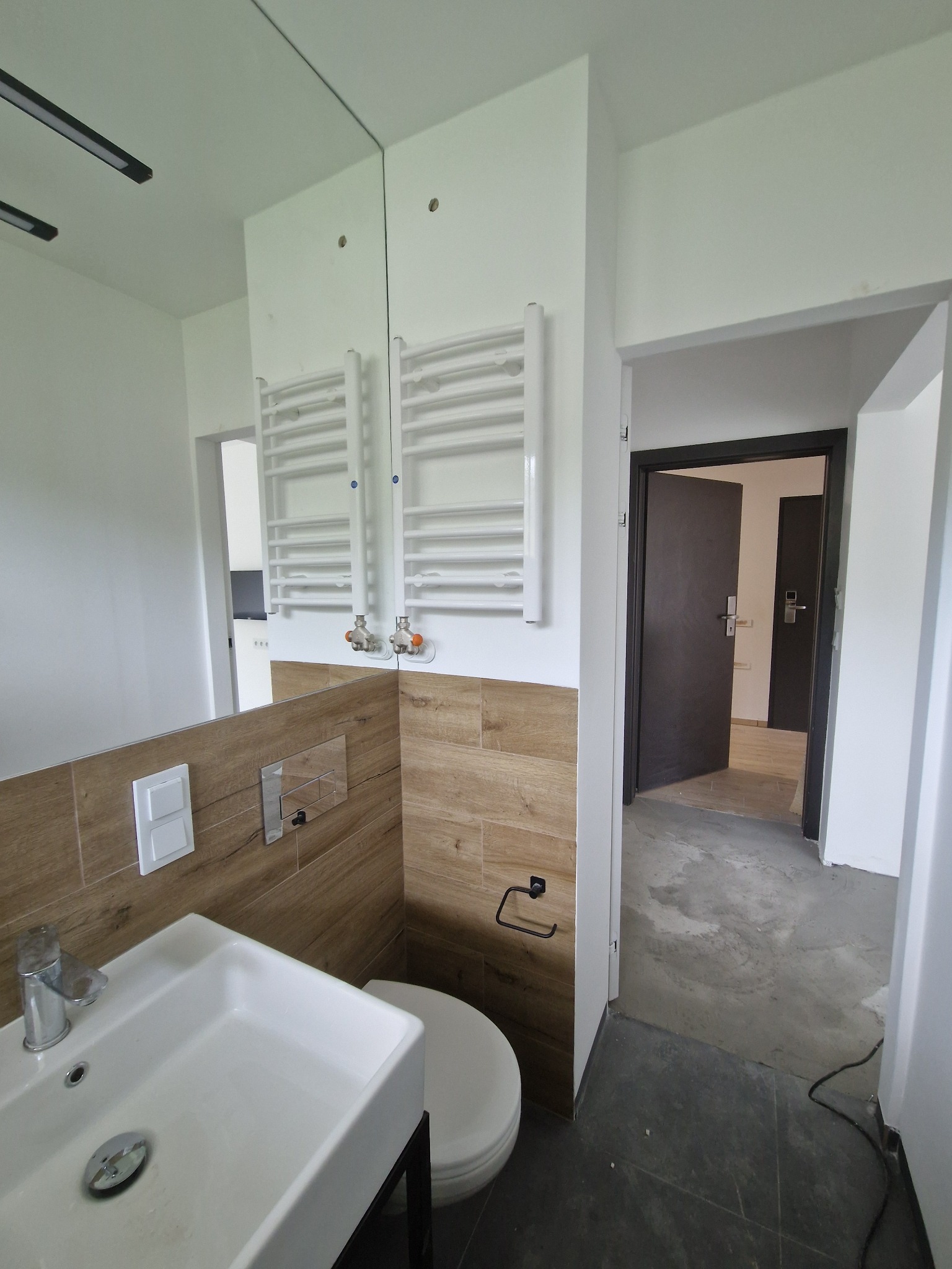 Wnętrze łazienki w trakcie wykańczania, widoczna umywalka, toaleta, grzejnik drabinkowy na ścianie z imitacją drewna, surowa podłoga i otwarte drzwi do kolejnego pomieszczenia.