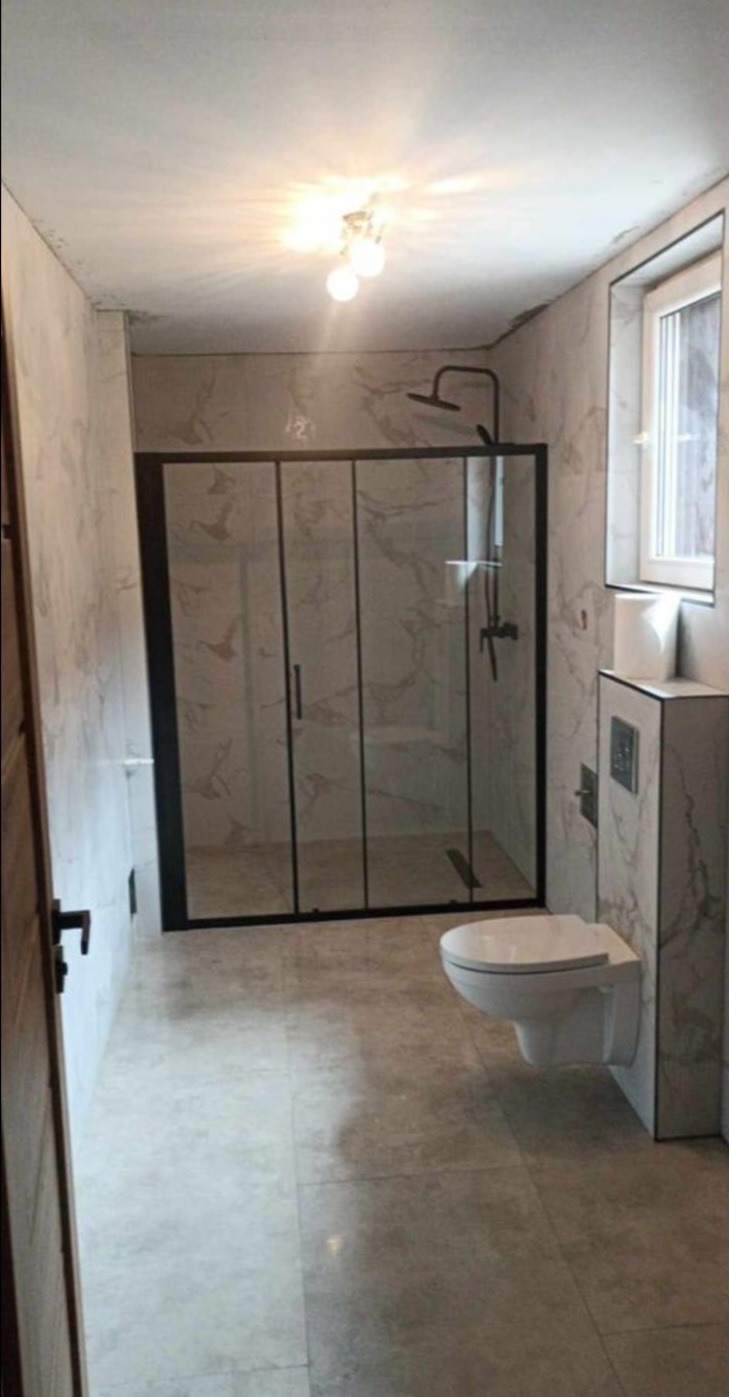 Wykończona łazienka z kabiną prysznicową w czarnej ramie, wiszącą toaletą i płytkami imitującymi marmur na ścianach i podłodze, oświetlona lampą sufitową z trzema żarówkami.