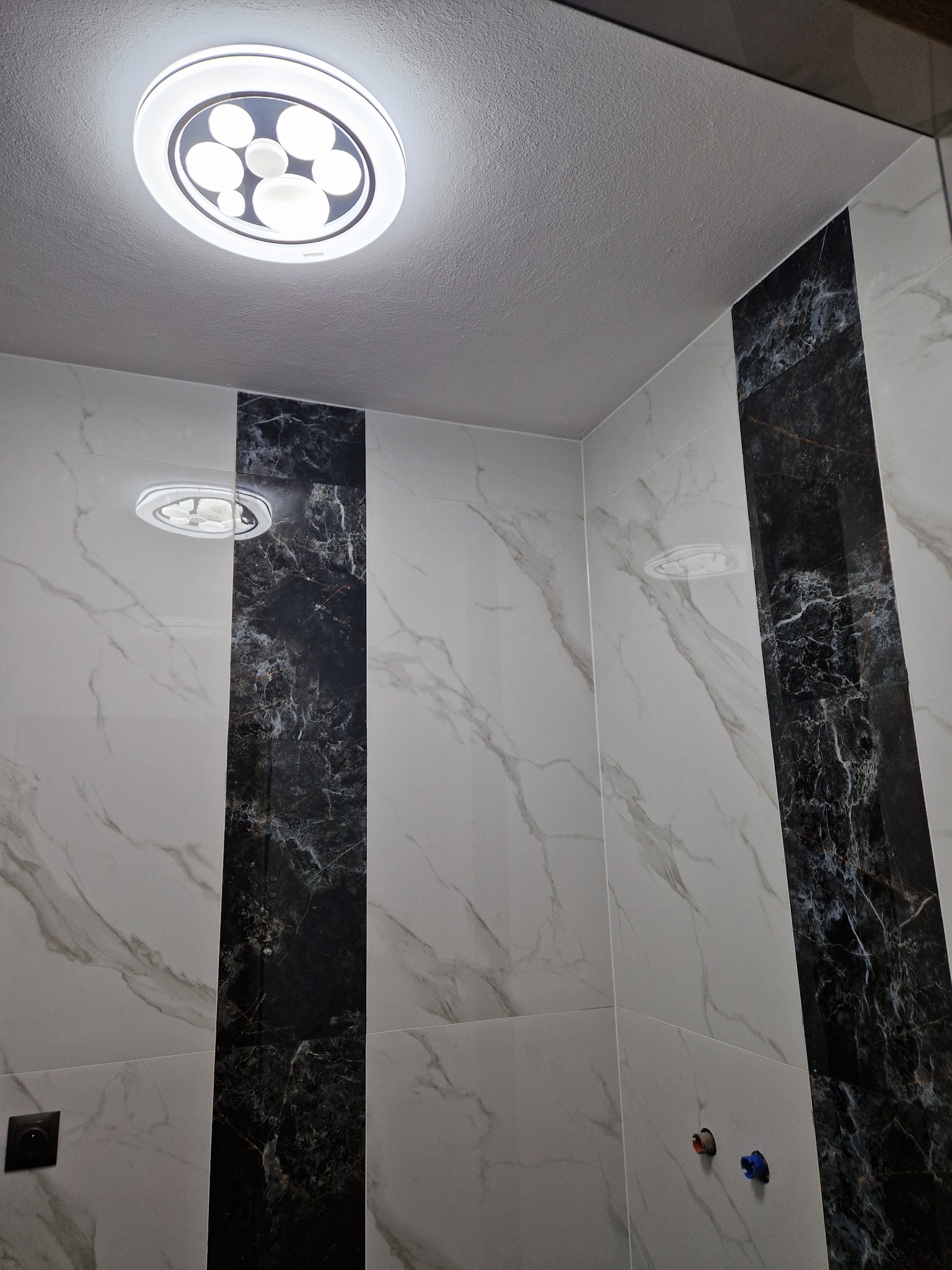 Ściany wyłożone białymi i czarnymi płytkami imitującymi marmur w łazience, z okrągłą lampą sufitową LED.