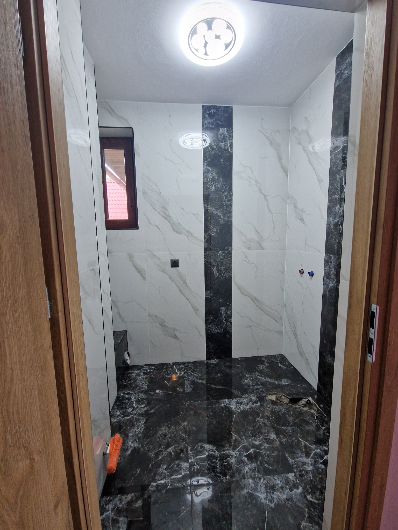 Wykończona łazienka z białymi i czarnymi marmurkowymi płytkami, widok na kabinę prysznicową z dwoma wystającymi zaworami, przez otwarte drewniane drzwi, z pomarańczowym narzędziem leżącym przy progu.