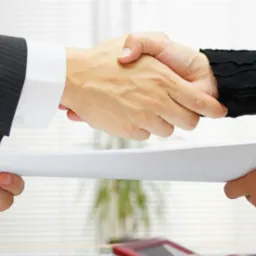 Uścisk dłoni nad dokumentem, w tle biurowe wnętrze z roletami i rośliną doniczkową.