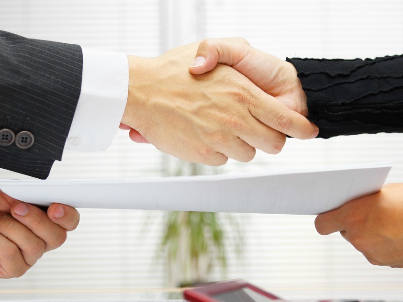 Uścisk dłoni nad dokumentem, w tle biurowe wnętrze z roletami i rośliną doniczkową.