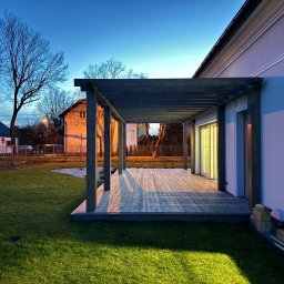 Ogród dla Ciebie Daniel Ositek - Nowoczesna pergola z drewnianą podłogą tarasową w Nowej Woli, oświetlona wieczornym światłem, widok na zadbany trawnik i fragment ogrodu z białym żwirem.