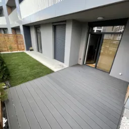 Nowoczesny taras z desek kompozytowych w odcieniach szarości, przylegający do budynku apartamentowego, z widokiem na trawnik z rolki i drewniane ogrodzenie.