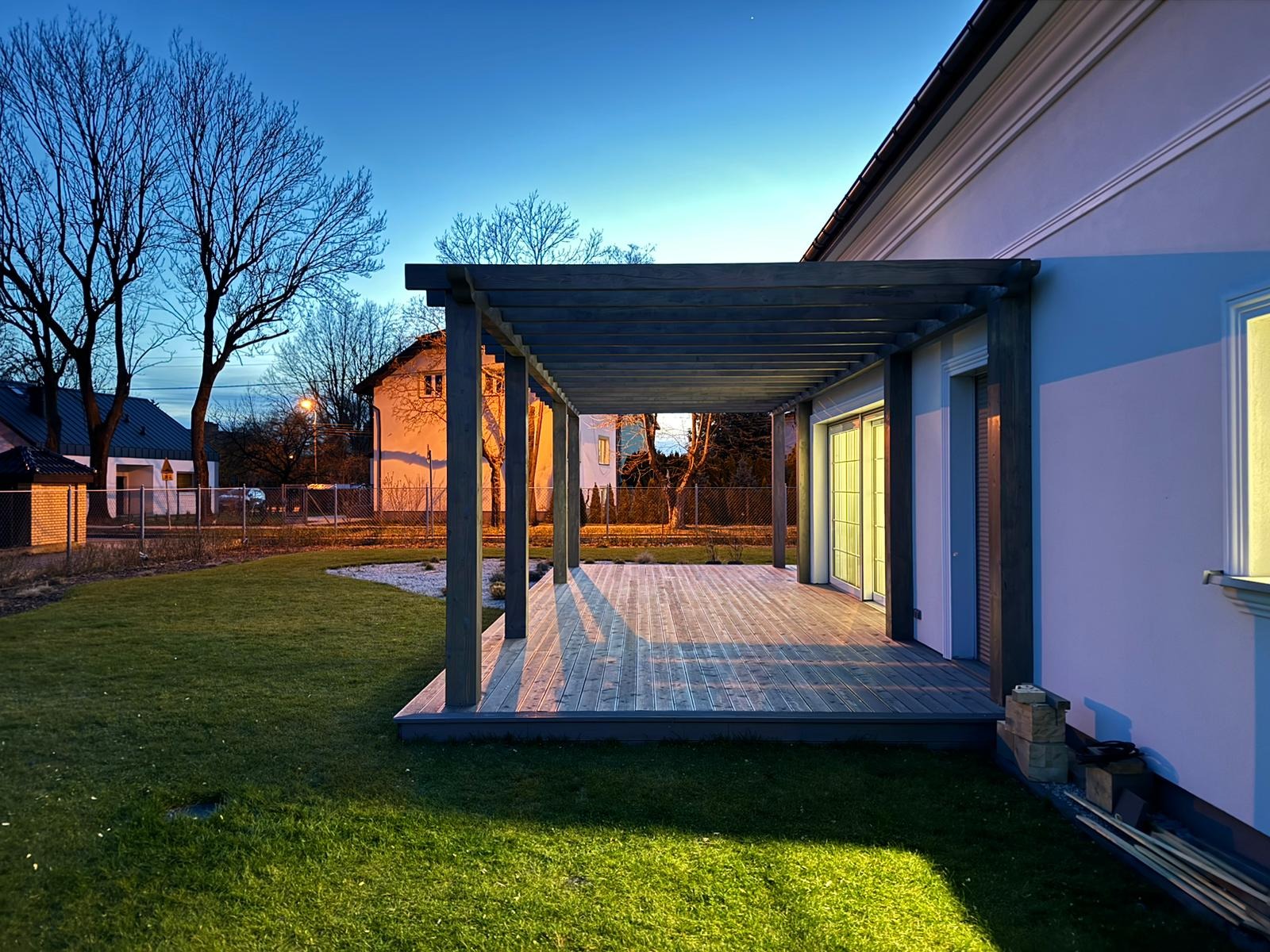 Nowoczesna pergola z drewnianą podłogą tarasową w Nowej Woli, oświetlona wieczornym światłem, widok na zadbany trawnik i fragment ogrodu z białym żwirem.