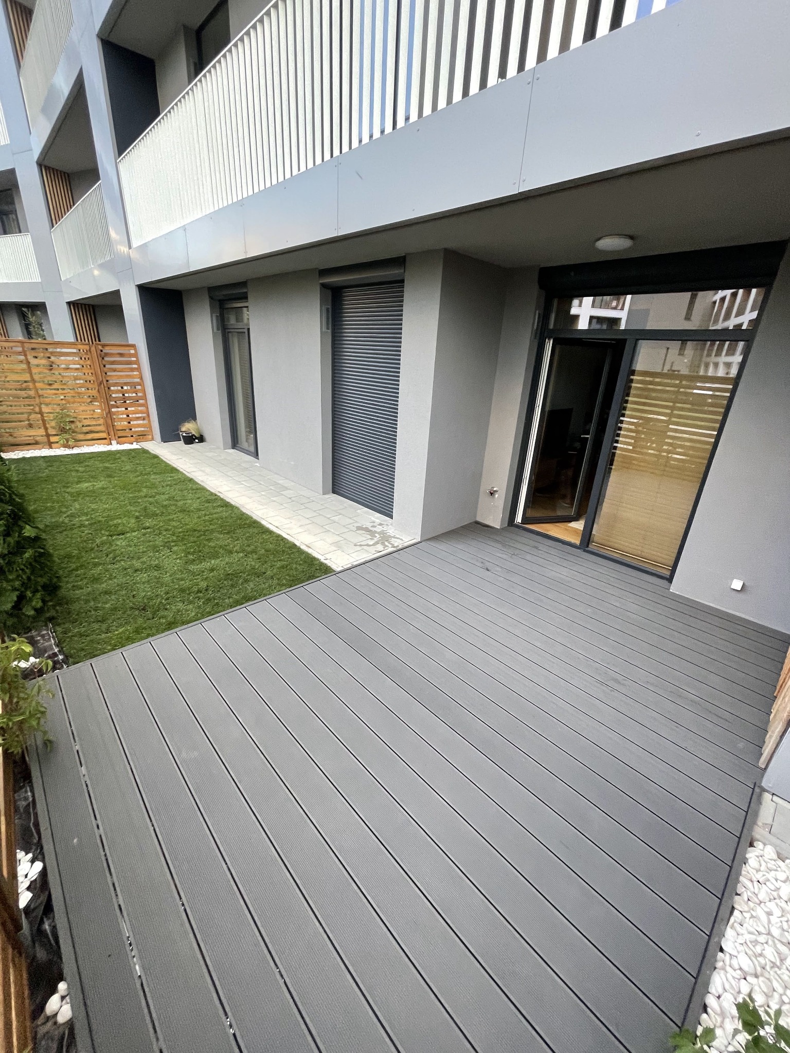 Nowoczesny taras z desek kompozytowych w odcieniach szarości, przylegający do budynku apartamentowego, z widokiem na trawnik z rolki i drewniane ogrodzenie.