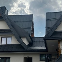 Usługi Blacharsko-Dekarskie Andrzej Kasperek - Widok na fragment dachu z blachy w kolorze grafitowym, z widocznymi obróbkami blacharskimi i kominem, na tle pochmurnego nieba. Na pierwszym planie fragment elewacji budynku z oknami i balkonem...