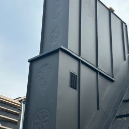 Usługi Blacharsko-Dekarskie Andrzej Kasperek - Czarny, metalowy komin z ozdobnymi tłoczeniami na dachu w trakcie prac dekarskich, widoczny fragment dachu z deskami i papą.
