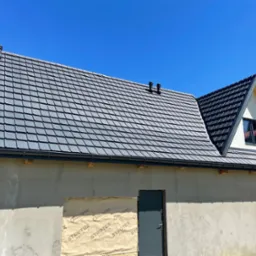 Nowy, grafitowy dach z dachówki ceramicznej z kominkami wentylacyjnymi na domu w trakcie wykończenia, z widocznym oknem na poddaszu i zabezpieczonym otworem drzwiowym.