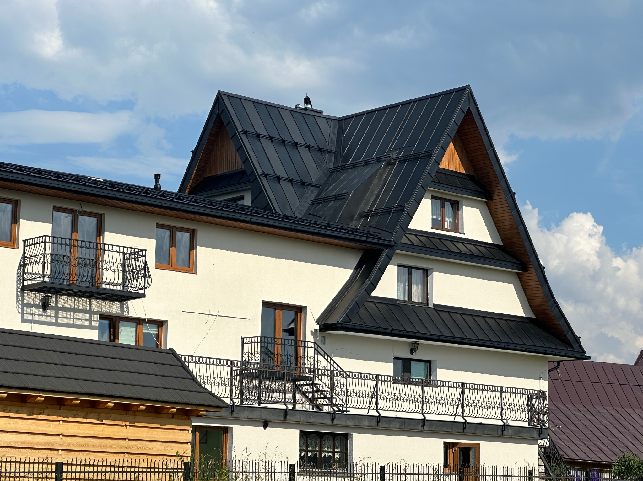 Nowoczesny dom z czarnym dachem z blachy na tle błękitnego nieba z chmurami. Widoczne detale architektoniczne, takie jak balkony z kutymi balustradami i drewniane wykończenia szczytów dachu.