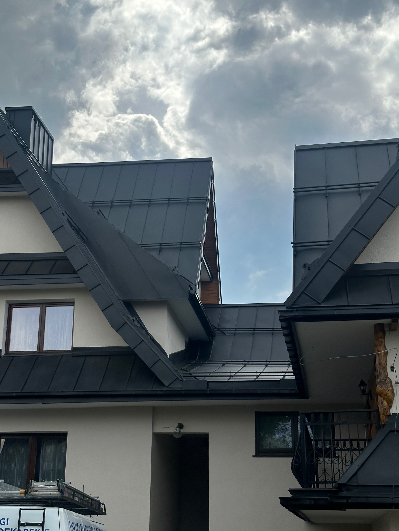 Widok na fragment dachu z blachy w kolorze grafitowym, z widocznymi obróbkami blacharskimi i kominem, na tle pochmurnego nieba. Na pierwszym planie fragment elewacji budynku z oknami i balkonem...