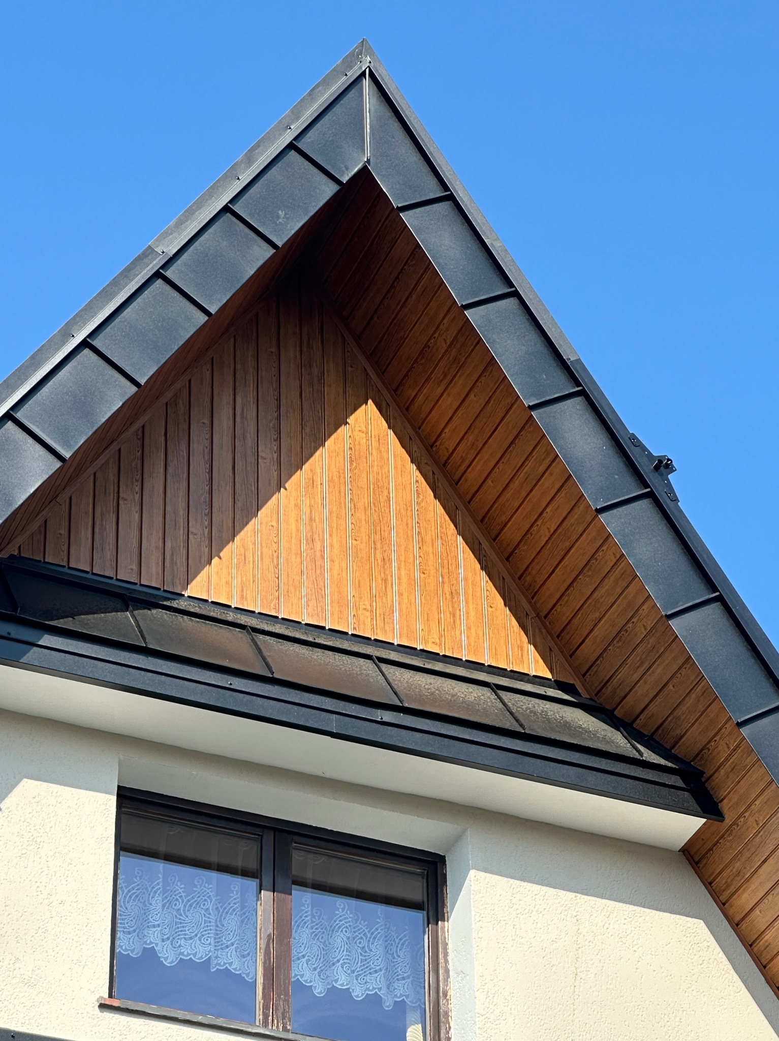 Szczyt dachu z czarnym kryciem blachodachówką panelową na rąbek stojący i boazerią drewnianą w pionowym układzie desek, widok z dołu na tle błękitnego nieba.