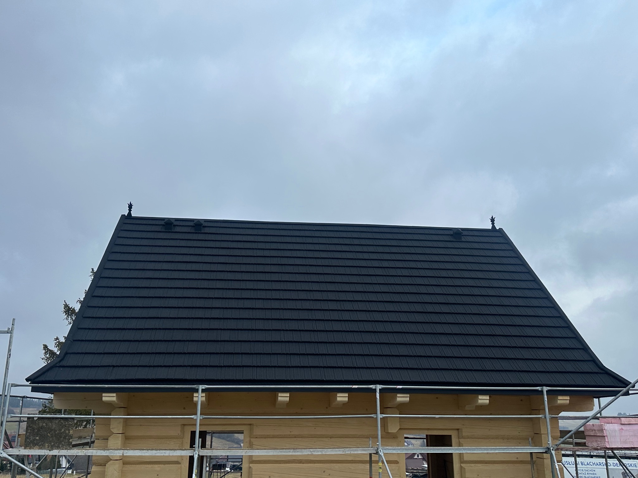 Nowy, czarny dach z metalowej blachy na drewnianym domu w stylu góralskim, widok z rusztowania, pochmurne niebo w tle.