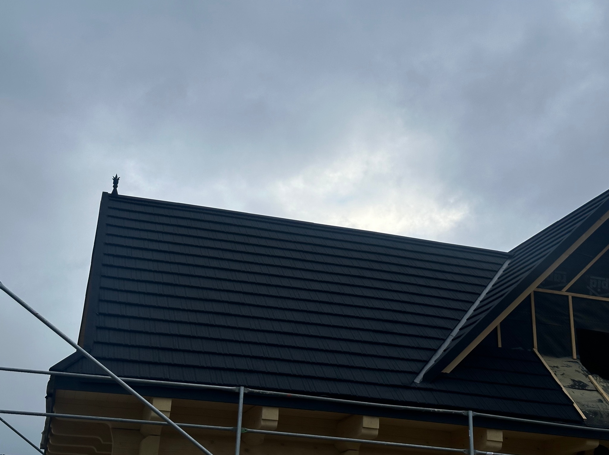 Nowy, ciemny dach z metalowej blachy na drewnianym domu w trakcie budowy, widoczny fragment rusztowania i ozdobny element na szczycie dachu.