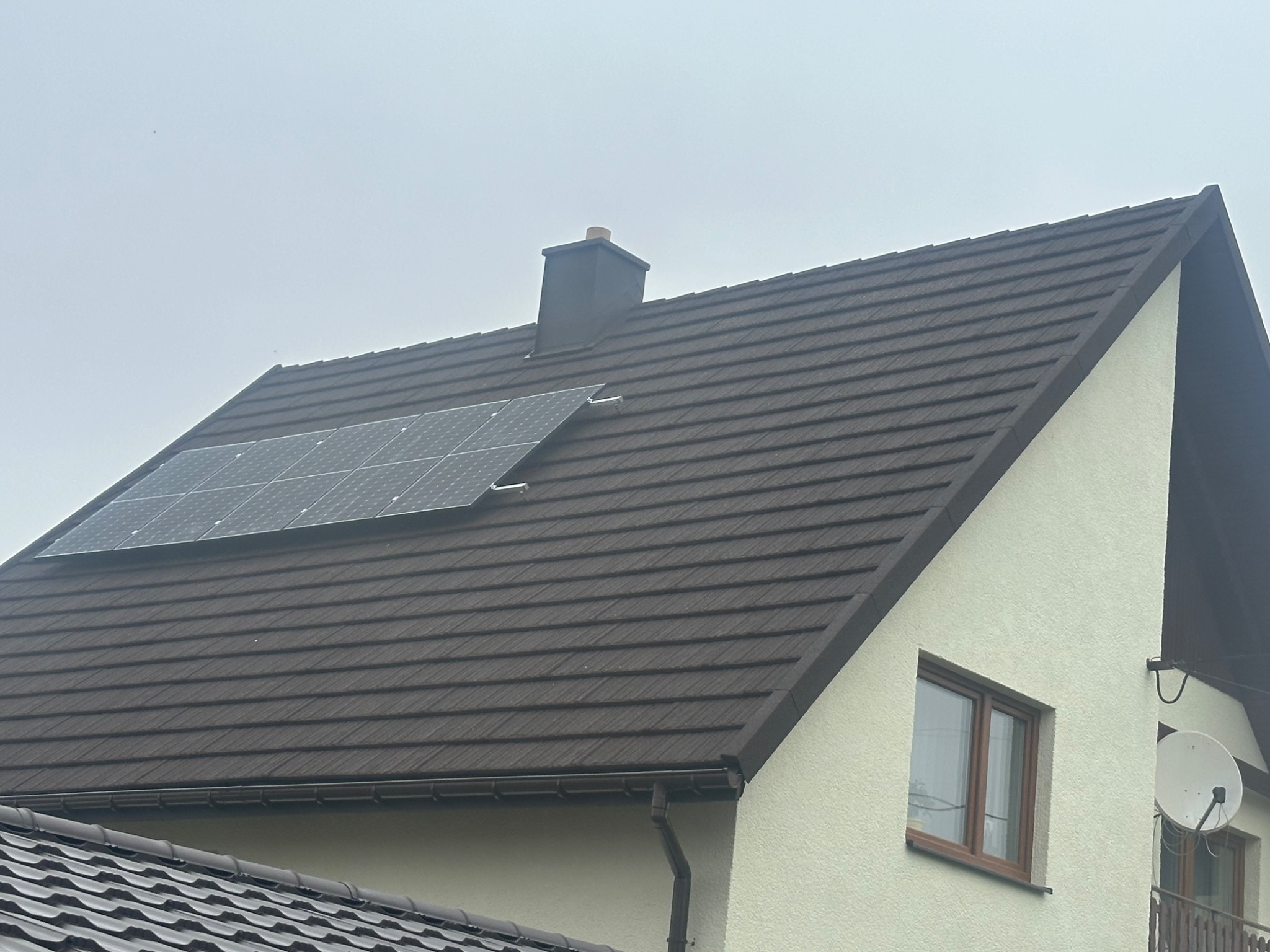 Brązowy dach z dachówki ceramicznej z zamontowanymi panelami słonecznymi, widoczny komin i rynna spustowa.