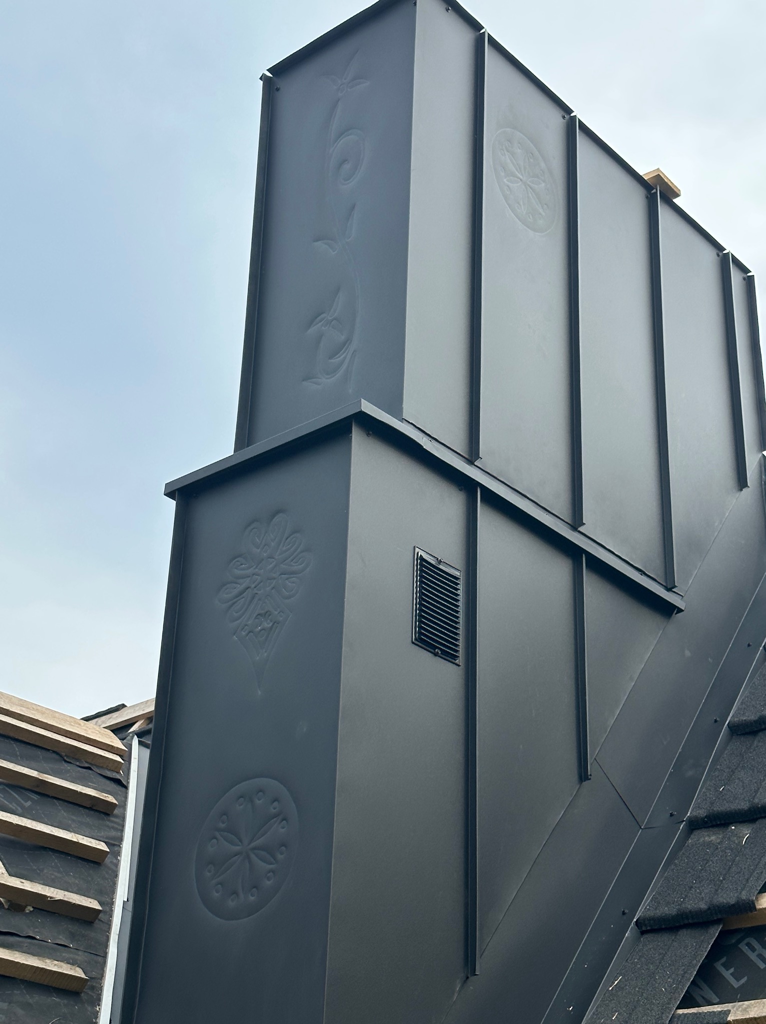 Czarny, metalowy komin z ozdobnymi tłoczeniami na dachu w trakcie prac dekarskich, widoczny fragment dachu z deskami i papą.