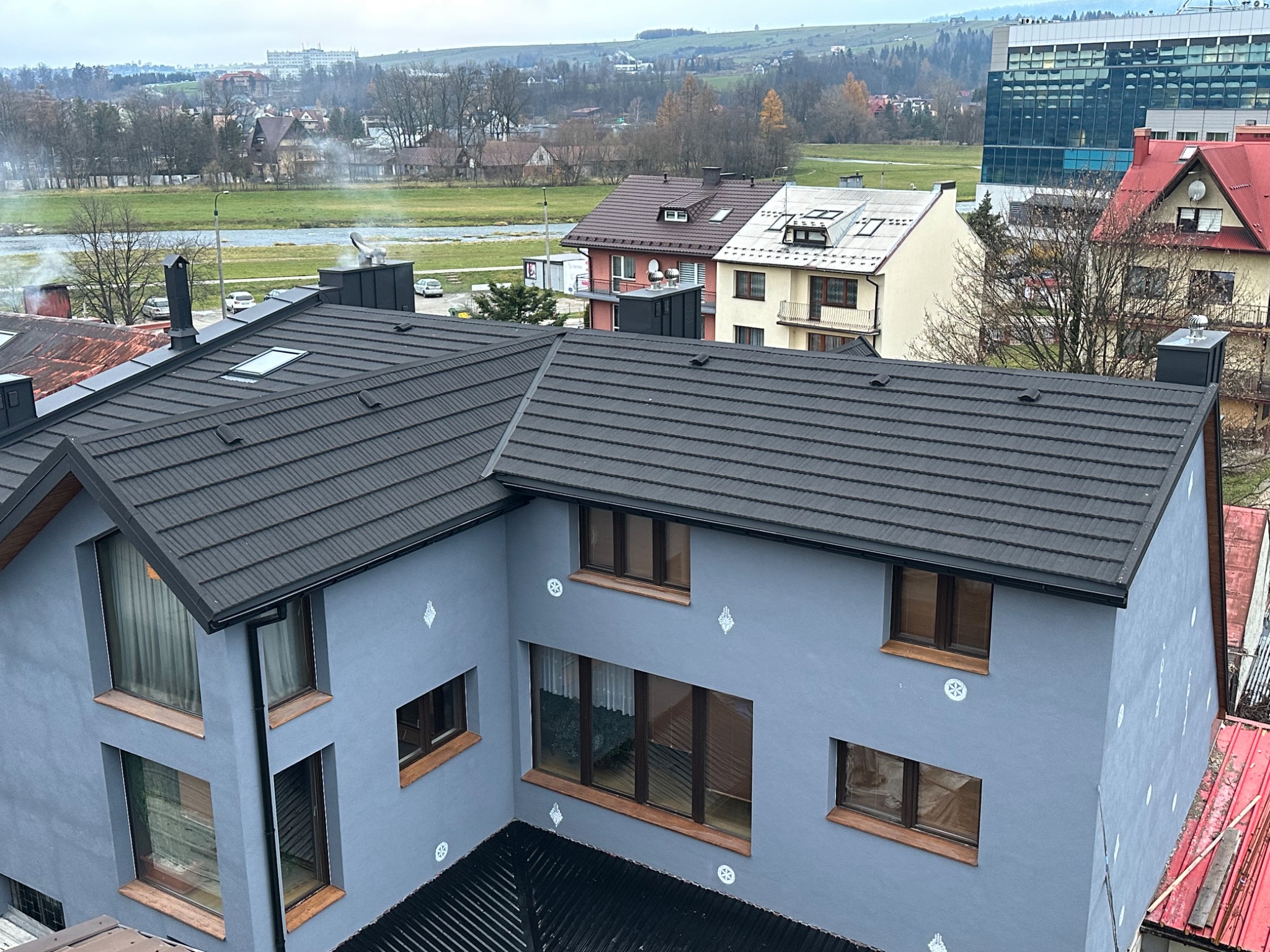 Nowoczesny, dwupiętrowy dom z ciemnoszarą dachówką, widoczny z góry, z oknami z brązowymi ramami i szarymi ścianami z dekoracyjnymi elementami, na tle zabudowy miejskiej i rzeki.
