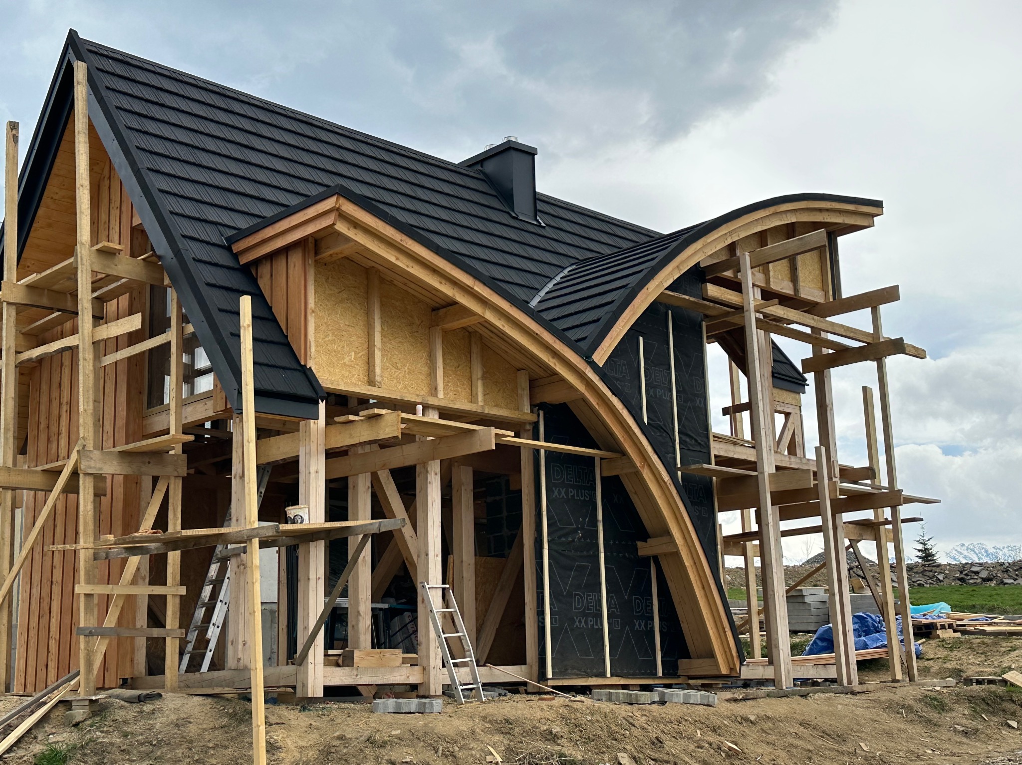 Dom w trakcie budowy z charakterystycznym, czarnym dachem z blachy na rąbek stojący, widoczne rusztowania i elementy konstrukcyjne z drewna, na tle pochmurnego nieba i gór.