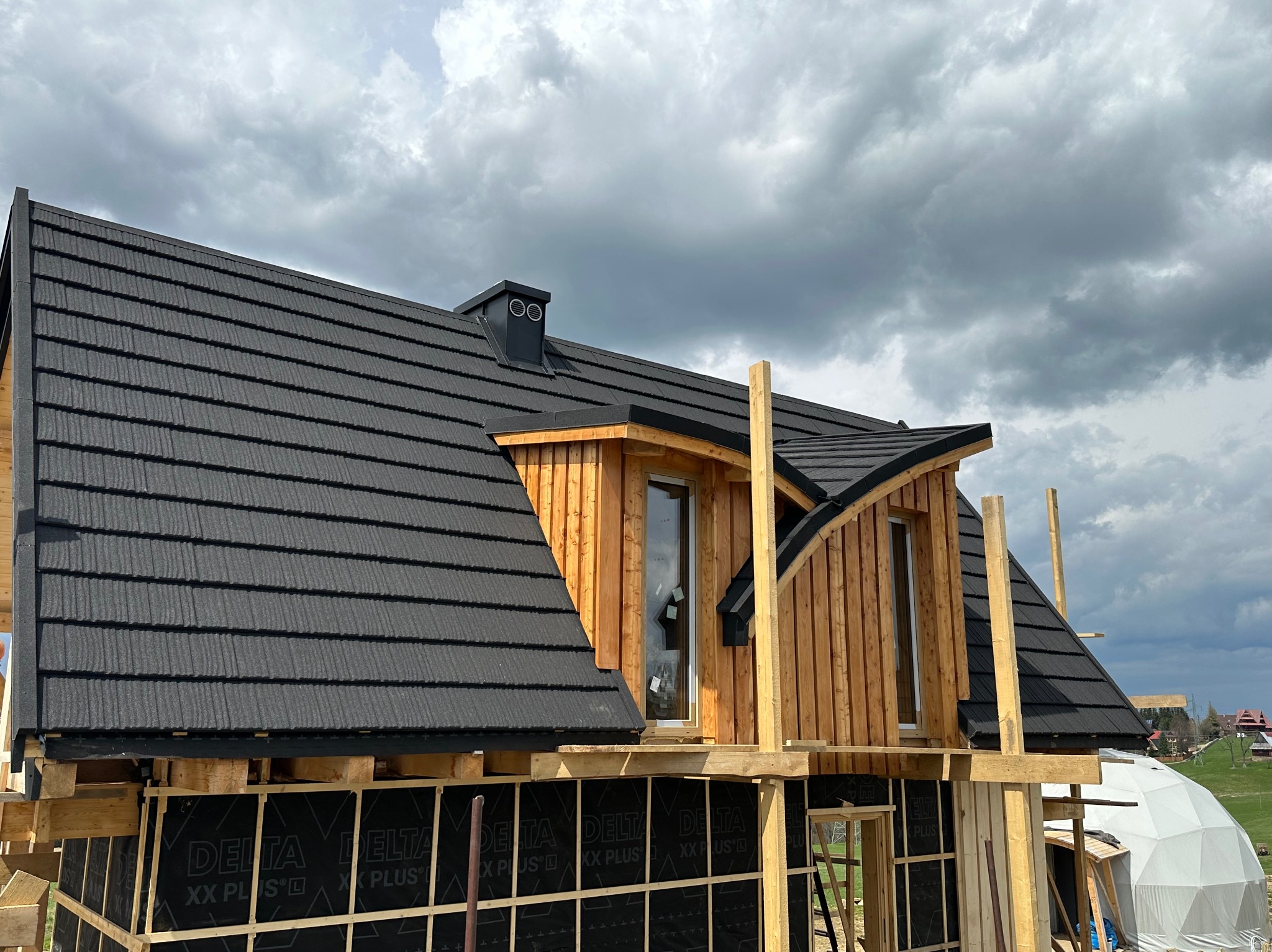 Nowy dom w trakcie budowy z czarnym dachem z blachodachówki modułowej, widoczne drewniane elementy konstrukcyjne, membrana dachowa Delta i fragment krajobrazu w tle pod pochmurnym niebem.