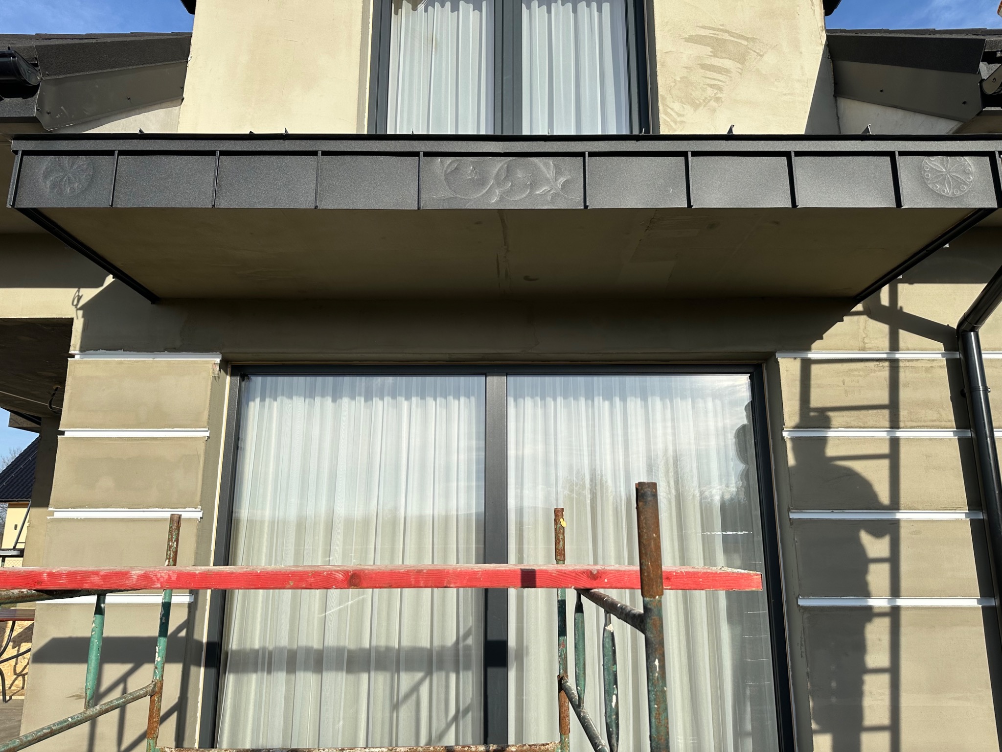 Szara obróbka blacharska nad oknem z delikatnym, wytłoczonym wzorem roślinnym i rozetą, widoczna z rusztowania. Ściana budynku w trakcie wykończenia, z widocznymi warstwami tynku i listwami.