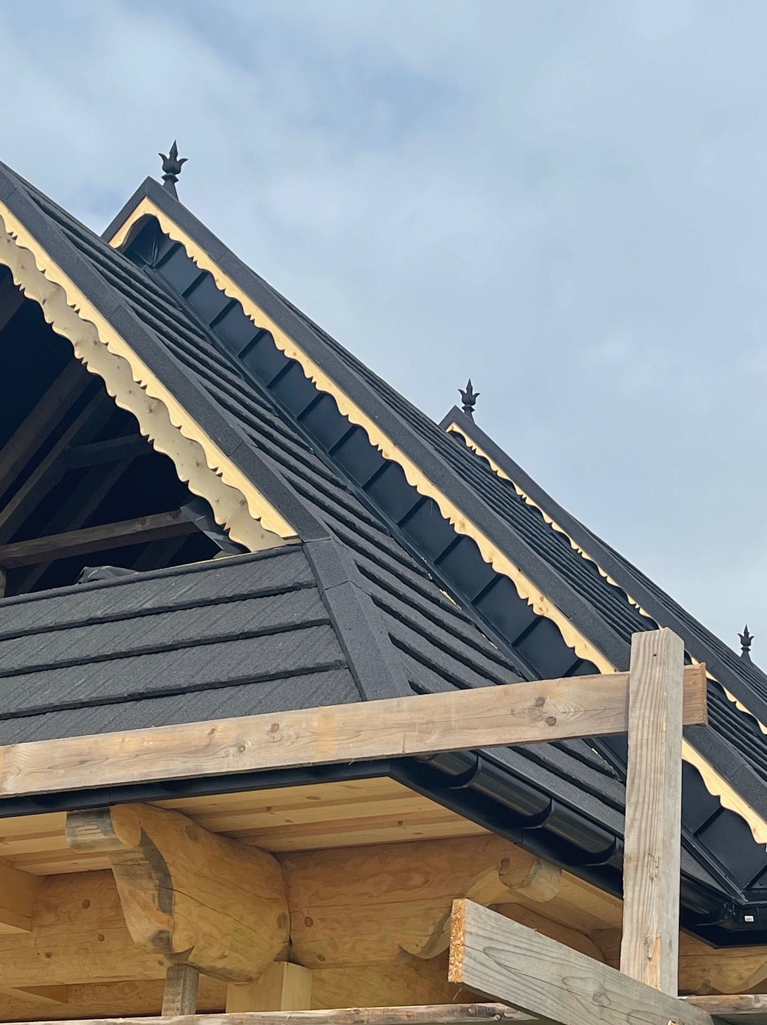 Fragment dachu z czarną dachówką bitumiczną i ozdobnymi elementami drewnianymi w kolorze jasnego drewna, zwieńczony metalowymi ozdobami, widoczne rusztowanie.
