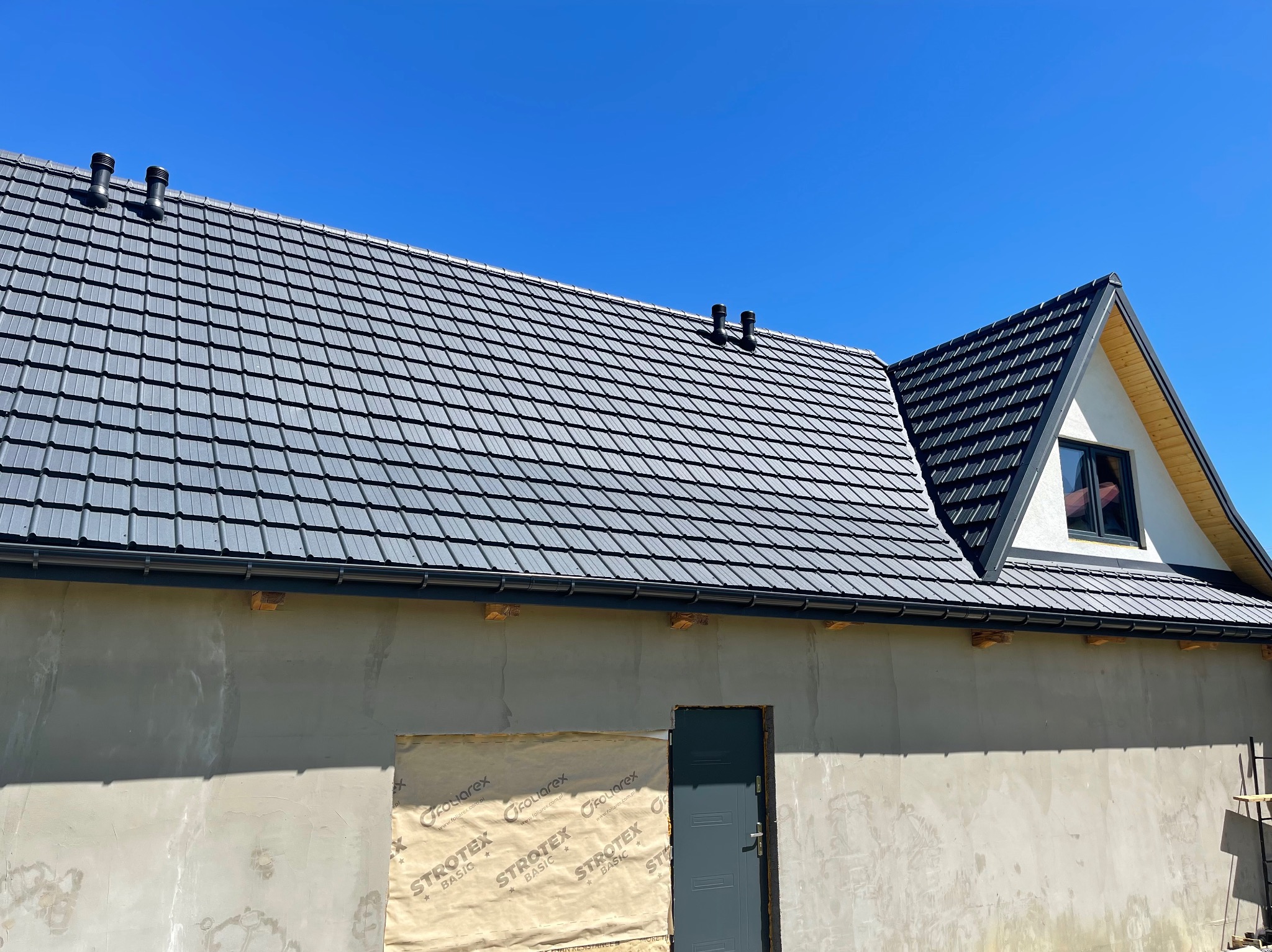 Nowy, grafitowy dach z dachówki ceramicznej z kominkami wentylacyjnymi na domu w trakcie wykończenia, z widocznym oknem na poddaszu i zabezpieczonym otworem drzwiowym.
