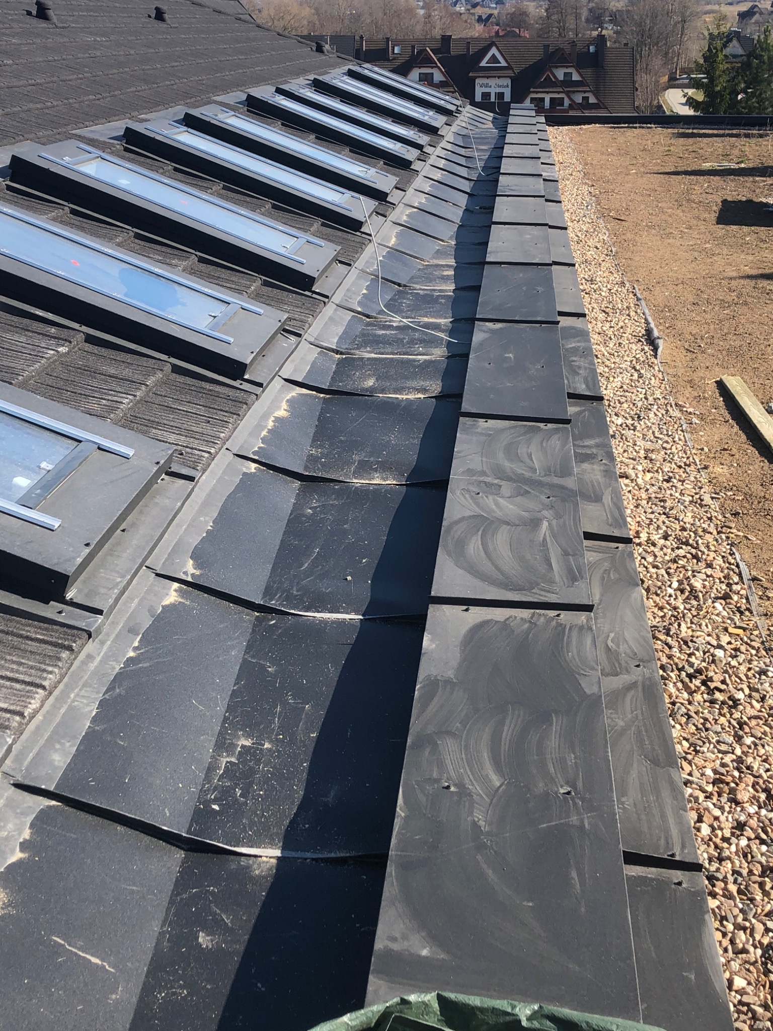 Fragment dachu z czarną dachówką, widoczne okna dachowe w rzędzie i obróbki blacharskie na krawędzi dachu, tło z widokiem na zabudowania.