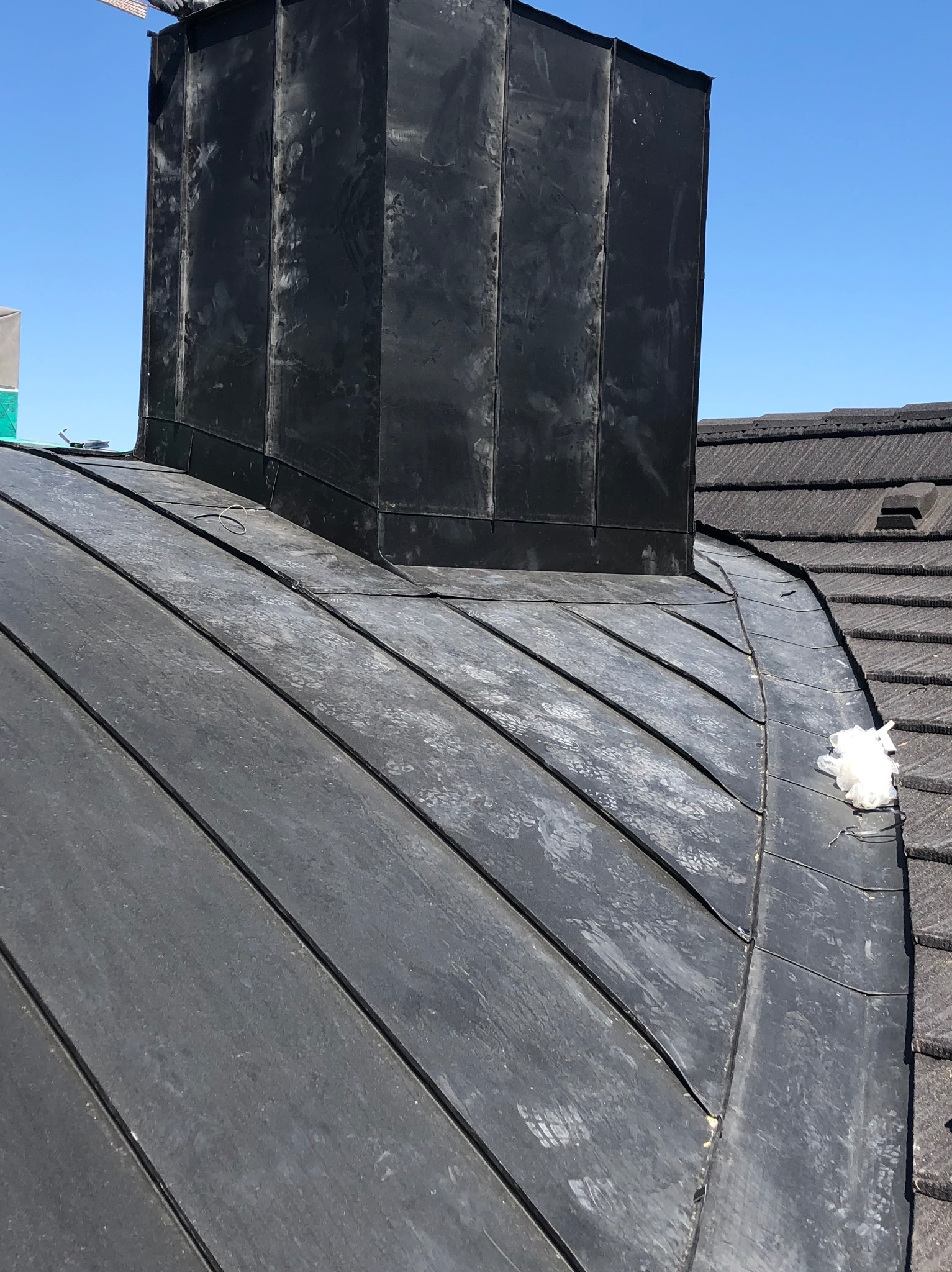 Szary dach z blachy na budynku w Szaflarach z widocznym kominem obłożonym blachą. Na dachu leży zwitek białego materiału.