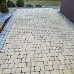 Myjnia mobilna - Ułożona kostka brukowa w Podegrodziu, widok z góry. Beżowa kostka z szarym obramowaniem, zielone krzewy w tle. Precyzyjne ułożenie, równa powierzchnia.