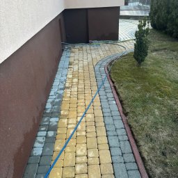 Myjnia mobilna - Częściowo umyta, dwukolorowa kostka brukowa (żółta i szara) przed budynkiem. Widoczny wąż ciśnieniowy i fragment trawnika z tujami. Efekt czyszczenia.