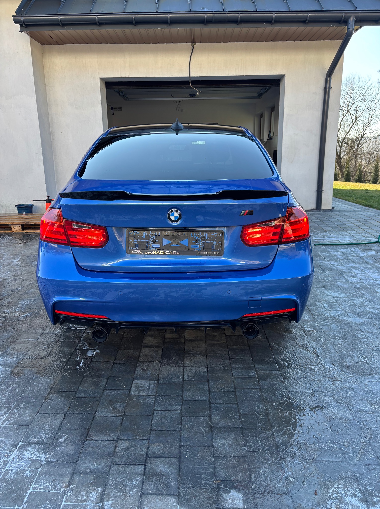 Błękitne BMW po myciu, widok z tyłu na mokrą kostkę brukową. Widoczne detale: światła, tablica rejestracyjna, logo BMW. W tle garaż i zieleń.