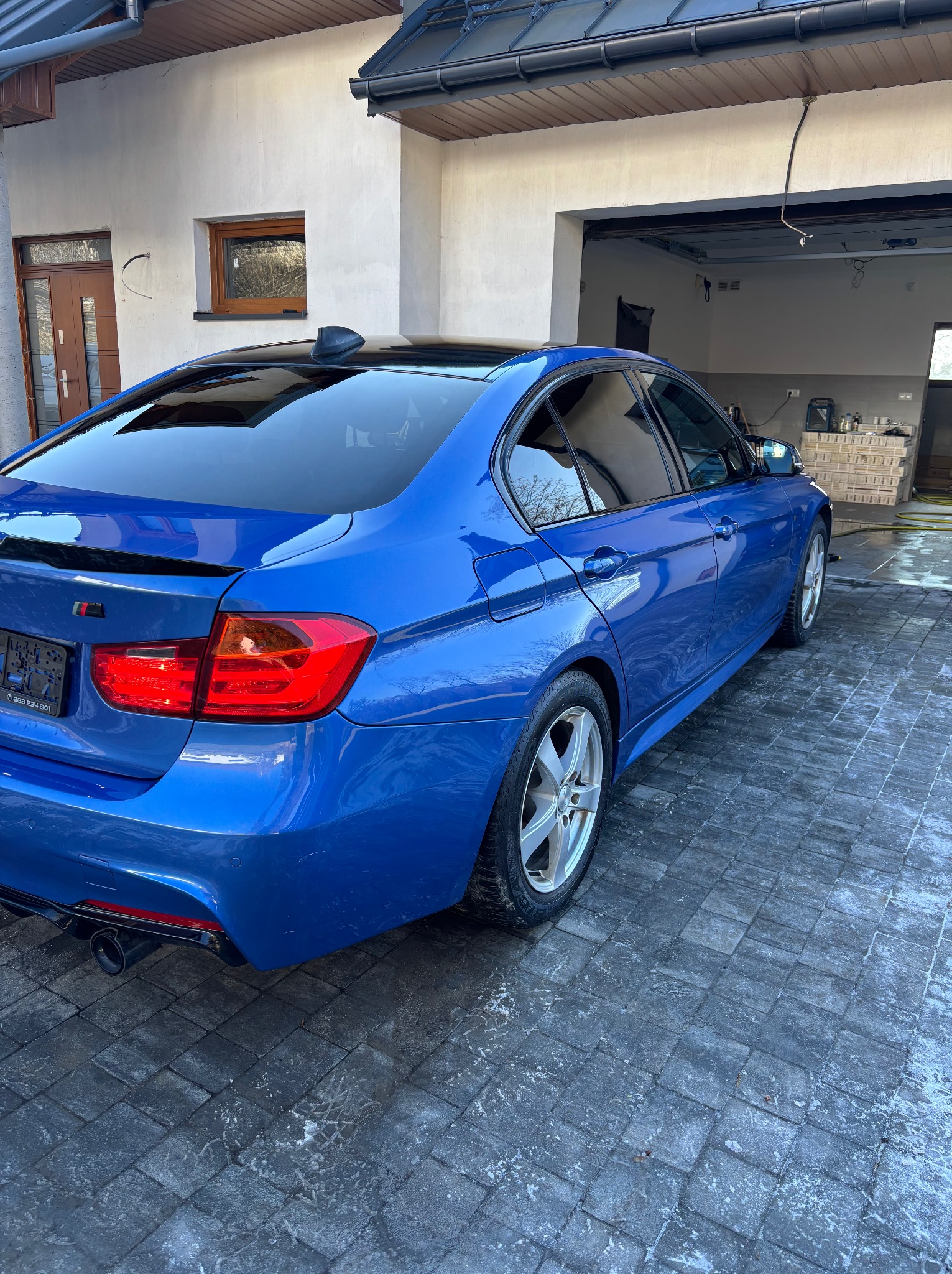 Błękitne BMW serii 3 po myciu, zaparkowane na mokrej kostce brukowej przed garażem. Widoczne ślady wody i odbicia na lakierze, podkreślające czystość.