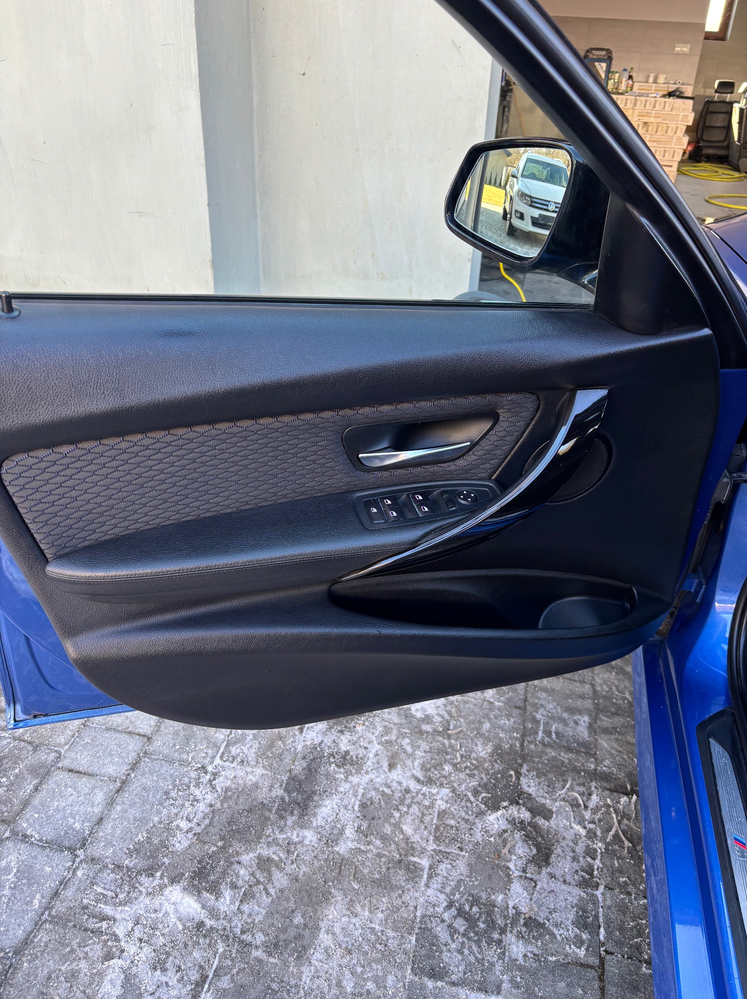 Wnętrze otwartych, błękitnych drzwi samochodu z widocznym lusterkiem bocznym, w którym odbija się biały van. Kostka brukowa pod drzwiami.