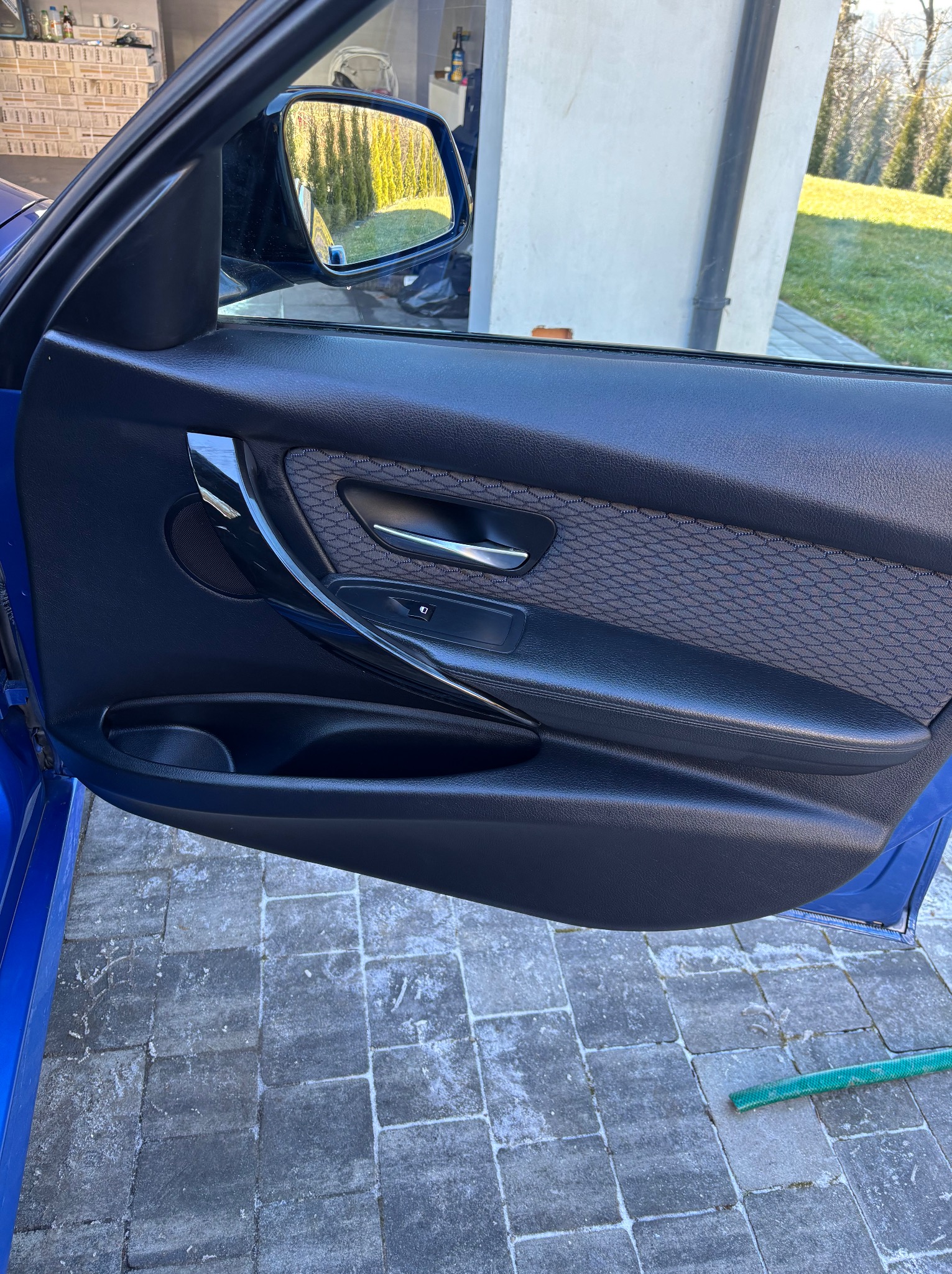 Wnętrze otwartych, błękitnych drzwi samochodu z czarną tapicerką i chromowanymi detalami. Widoczny fragment kostki brukowej i węża na zewnątrz.