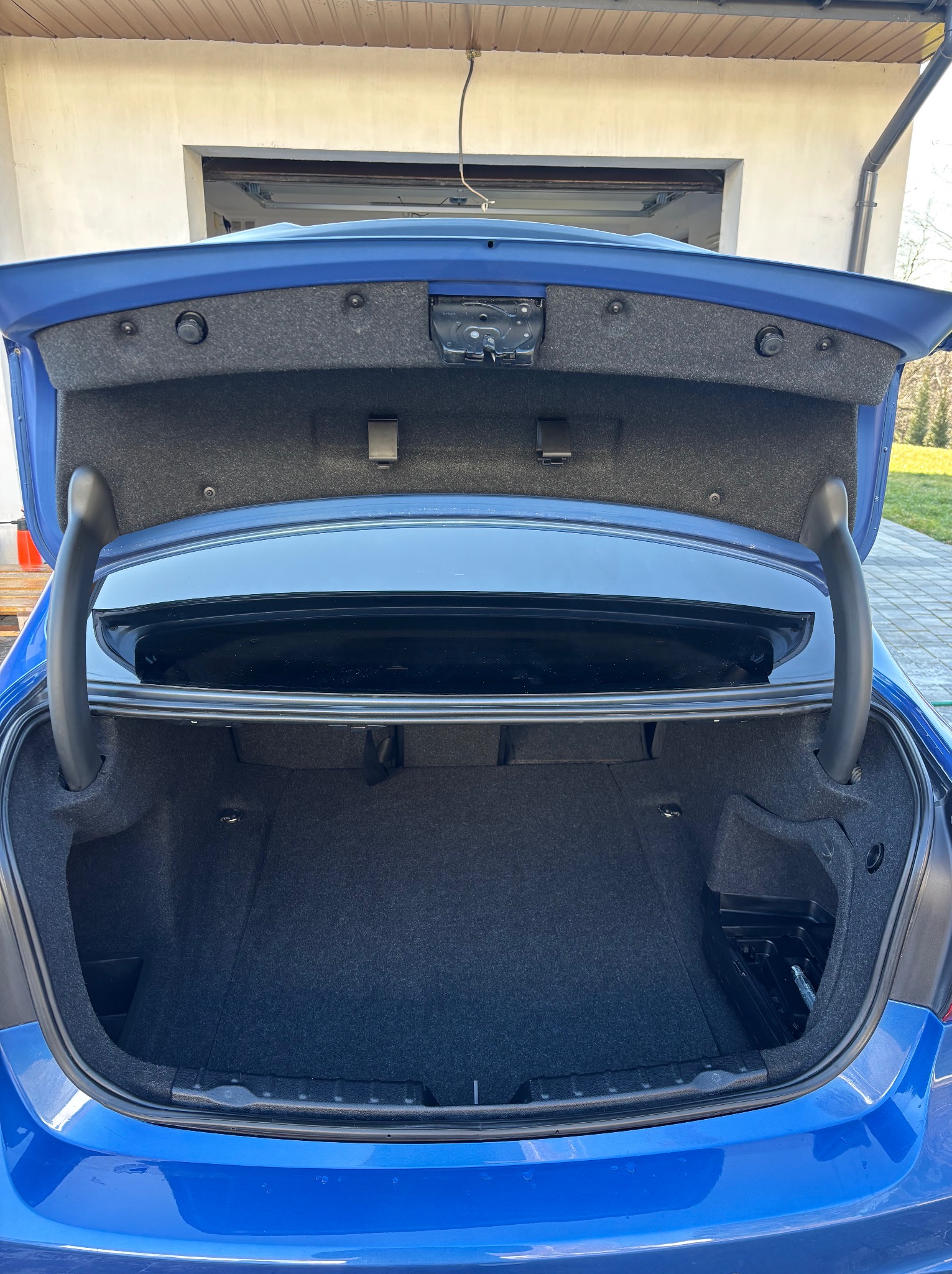 Otwarty bagażnik niebieskiego samochodu osobowego z widocznym wnętrzem, elementami mocującymi i czarną wykładziną. W tle brama garażowa i zieleń.