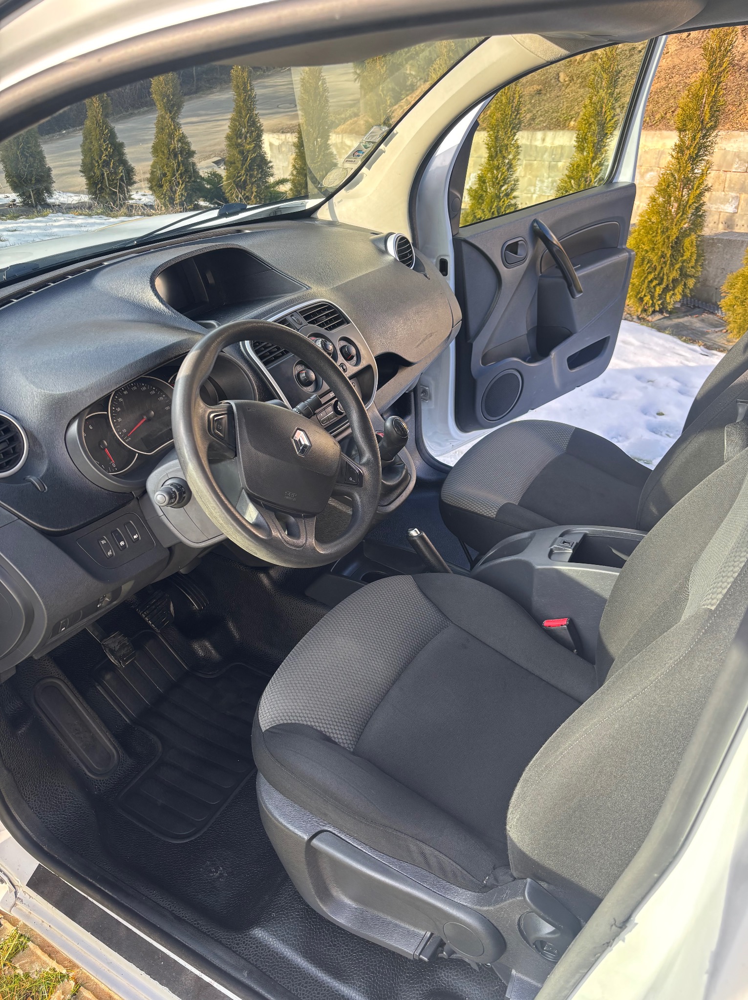 Wnętrze białego Renault Kangoo po czyszczeniu: czarne dywaniki, szare fotele, deska rozdzielcza. Widok z otwartymi drzwiami na zewnątrz z zielonymi tujami i śniegiem.