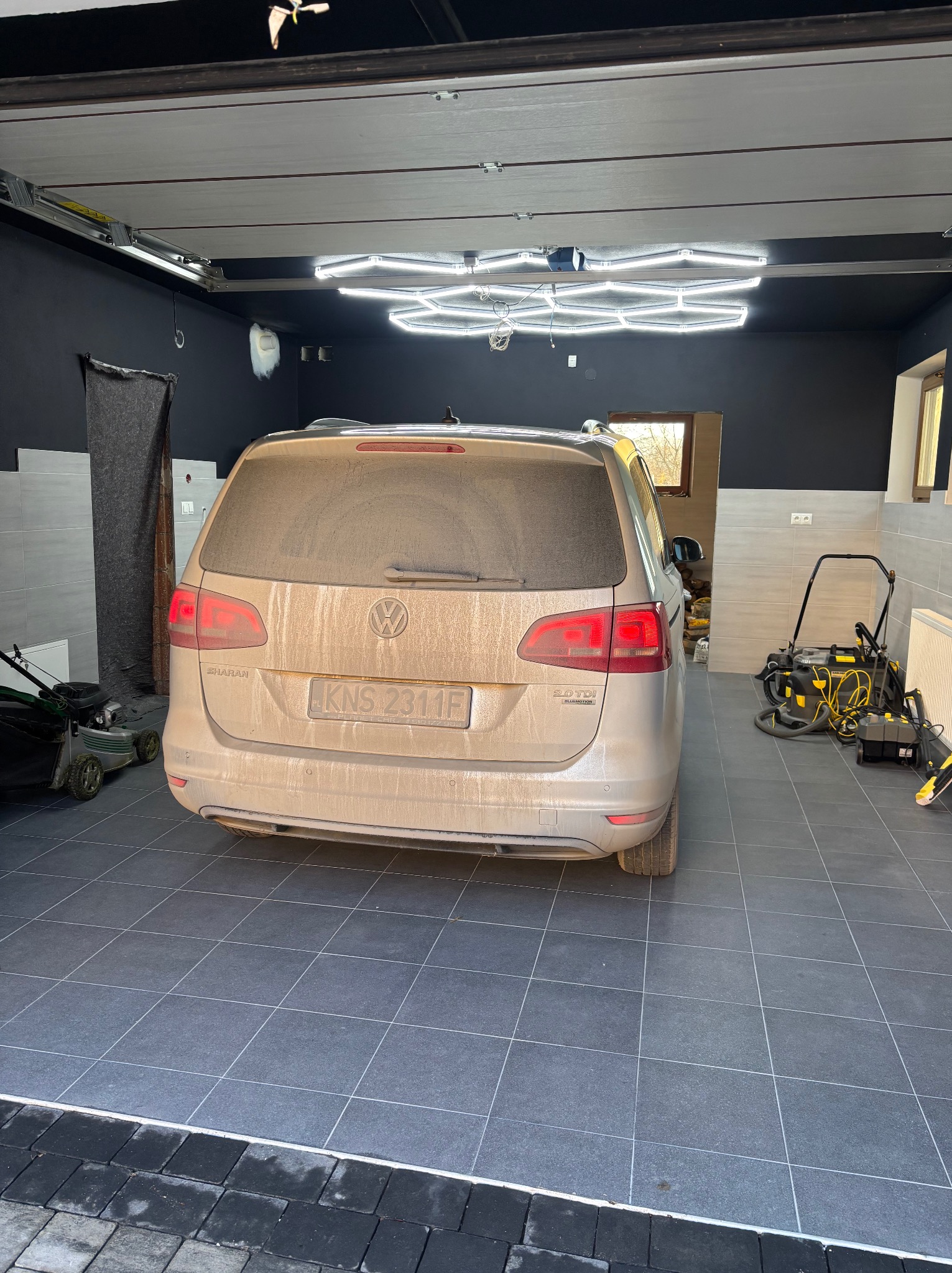 Bardzo brudny, beżowy van VW Sharan w garażu z szarą podłogą z płytek. W tle widoczny sprzęt do mycia ciśnieniowego i kosiarka. Oświetlenie garażowe LED w kształcie plastra miodu.