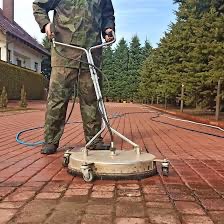Czyszczenie kostki brukowej myjką ciśnieniową z okrągłą dyszą. Mężczyzna w kamuflażu obsługuje urządzenie na tle domu i drzew.