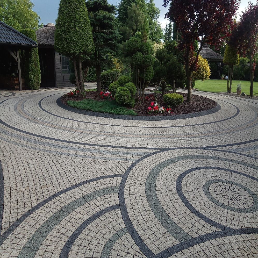 Okrągły podjazd z kostki brukowej ułożonej we wzory, otoczony zielenią i drzewami. Widoczne altany z krytą strzechą w tle. Estetyczne wykończenie posesji.